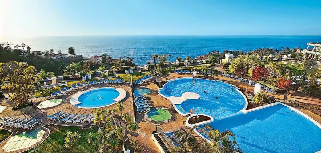 A large resort with terraced gardens, sun loungers, and umbrellas overlooking the ocean and distant mountains under a clear sky.