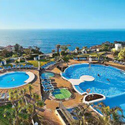 A large resort with terraced gardens, sun loungers, and umbrellas overlooking the ocean and distant mountains under a clear sky.