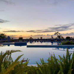 A large resort with terraced gardens, sun loungers, and umbrellas overlooking the ocean and distant mountains under a clear sky.