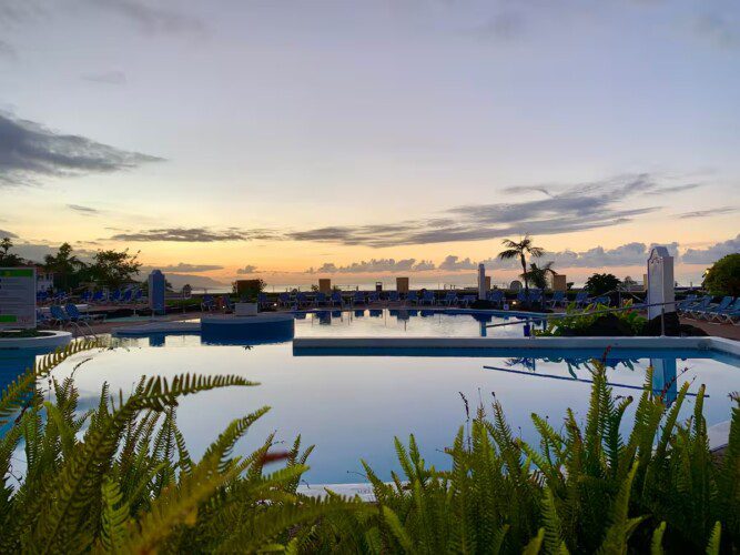A large resort with terraced gardens, sun loungers, and umbrellas overlooking the ocean and distant mountains under a clear sky.