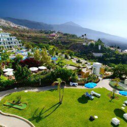 A large resort with terraced gardens, sun loungers, and umbrellas overlooking the ocean and distant mountains under a clear sky.