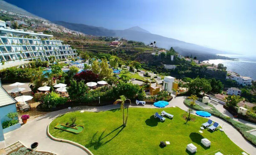 A large resort with terraced gardens, sun loungers, and umbrellas overlooking the ocean and distant mountains under a clear sky.
