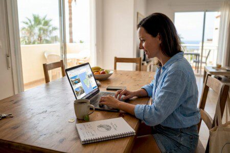 Eine Frau sitzt am Tisch und stöbert am Laptop nach passenden Ferienwohnungen.